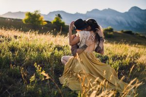 intimate couple embraces while woman's yellow dress train flows down the grassy hill in the colorado mountains