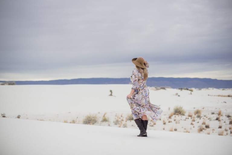 a young blonde woman stands in White Sands New Mexico looking out at the horizon with a long dress, tan hat and brown boots