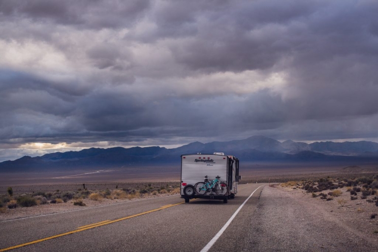 An RV towing a Springdale mini travel trailer driving along a desert road in Nevada into a storm of dark blue clouds with mountains in the distance