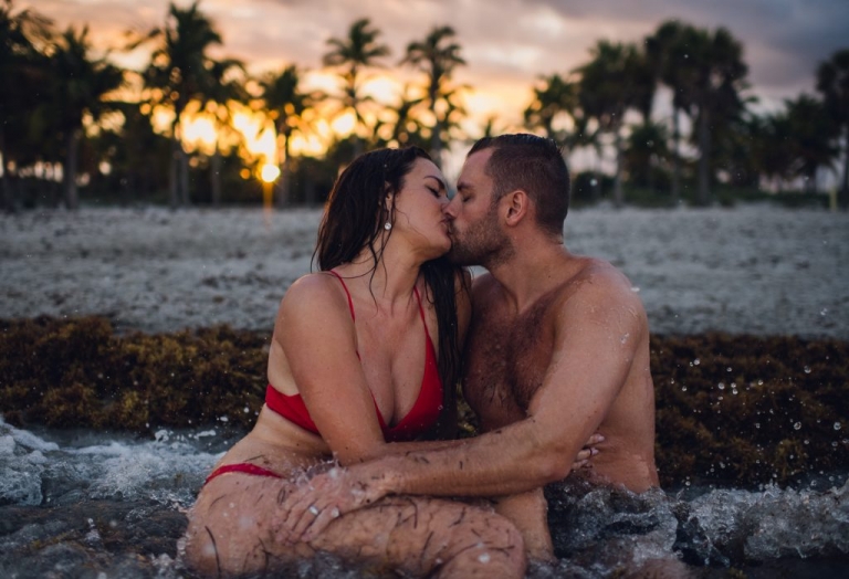 A couple shares a passionate kiss while sitting in the waves on a tropical beach at sunset