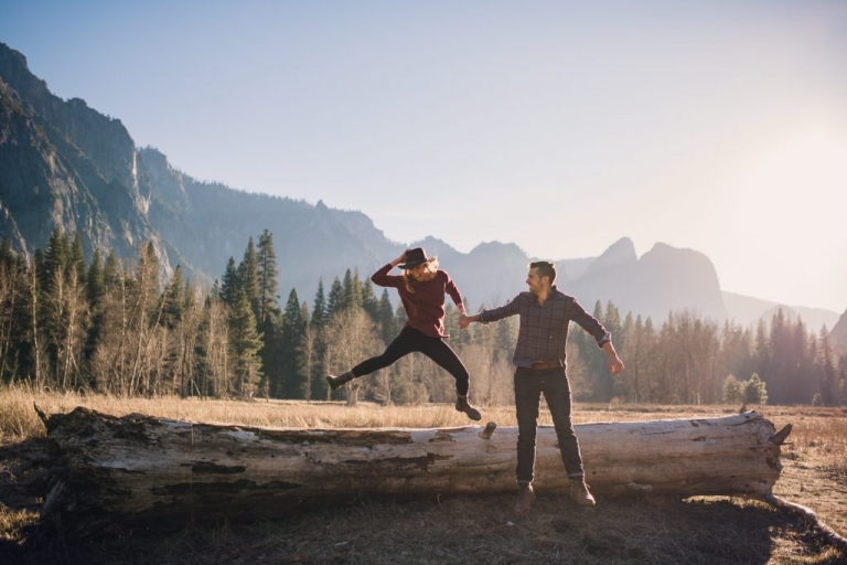 Adventurous couples jumps off of a fallen tree in Yosemite Valley while the woman holds her hat on her head