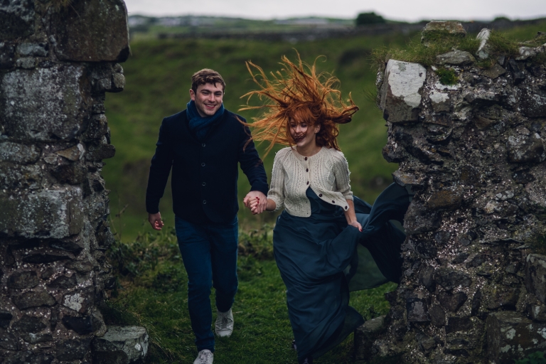 young couple walking through rocky remains of a castle in northern ireland as the woman's red hair blows wildly in the wind