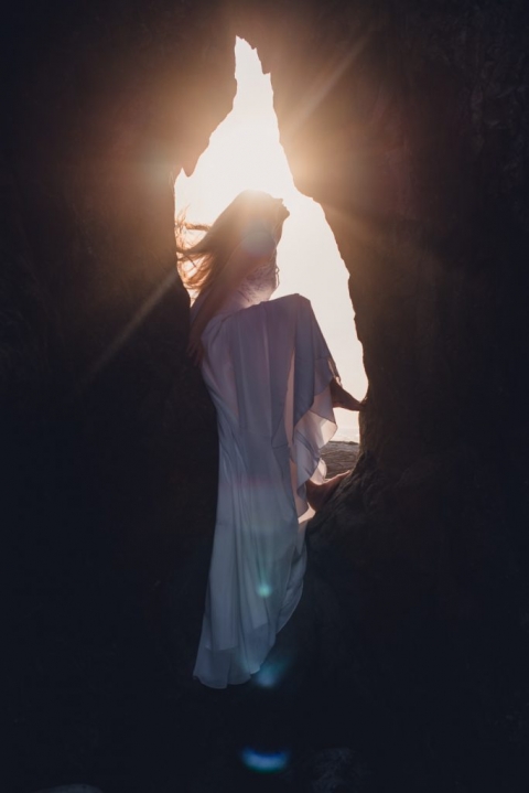 silhouette of a woman in a wedding dress sitting in the opening of a rock whipping her hair back with the sun flaring through