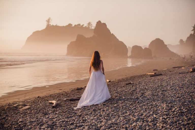 red hair bride with train walking along a rocky beach at sunset with seastacks in the distance