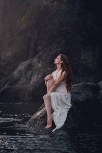 red head woman in a white dress sitting on a rock hugging her knee to her chest and gazing at the sunset