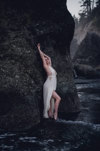 woman in a white dress leaning against a seastack on ruby beach stretching towards the sky