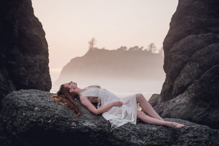 woman in a white dress laying down on a rock formation gazing at the sky