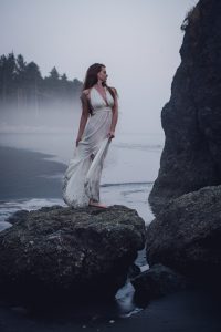 woman in a white dress standing on a rock at ruby beach after sunset with mist rolling in from the sea