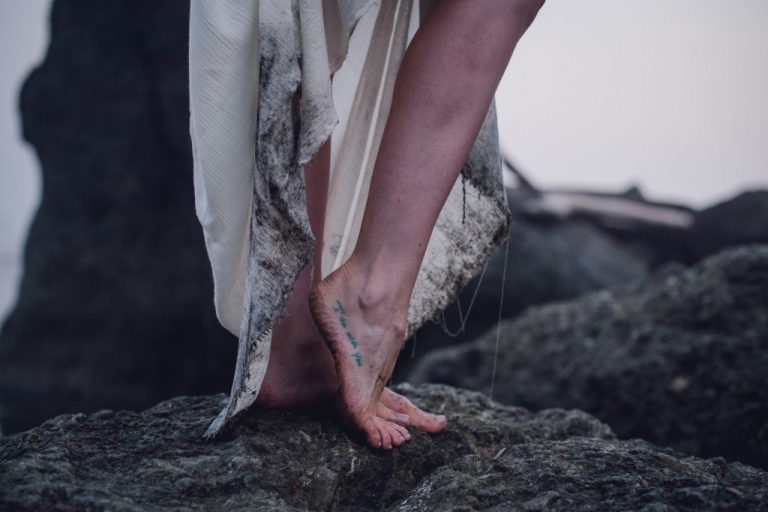 woman's leg with dark sand on the hem of her white dress showing off a foot tattoo that reads "i am with you"