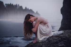 woman in a white dress with long red hair hugging her knees to her chest while sitting on a rock on a misty beach