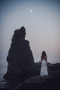 woman in a white dress staring at the moon next to a rock formation on ruby beach washington