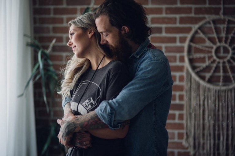 inked man hugs blonde woman from behind while they gaze out the window of their seattle loft 