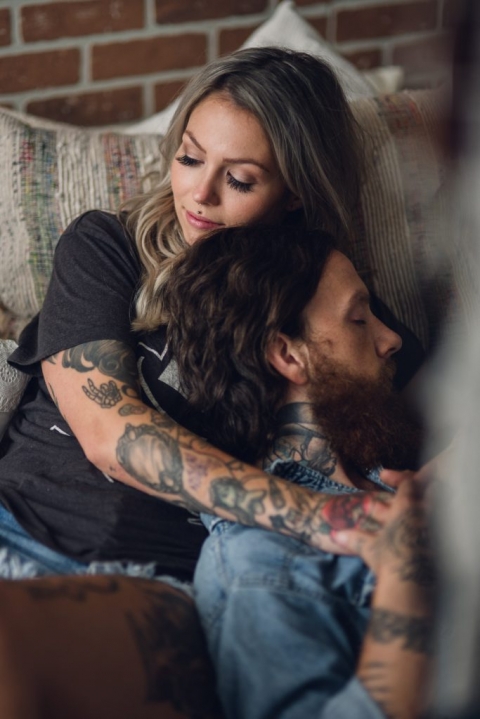 blonde woman with a tattoo sleeve embraces her bearded partner while relaxing at home