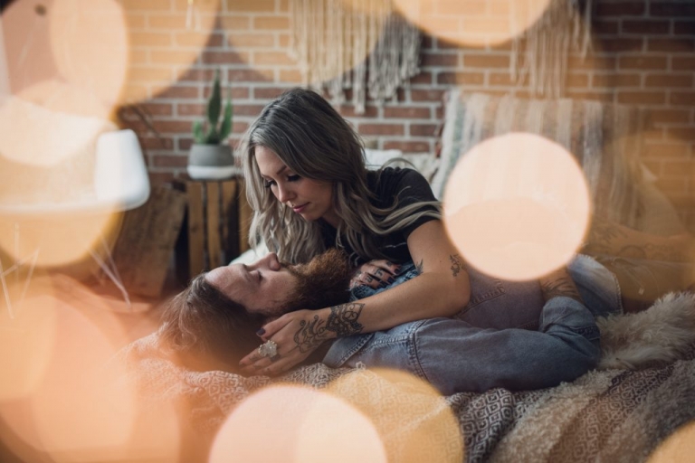 tattooed couple lies facing each other on a bed with the glow of lights around them